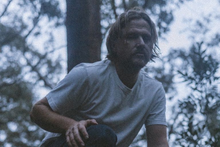 Promo photo of Simon Finch from Australian band The Tambourine Girls crouching outdoors in a white t-shirt surrounded by trees.